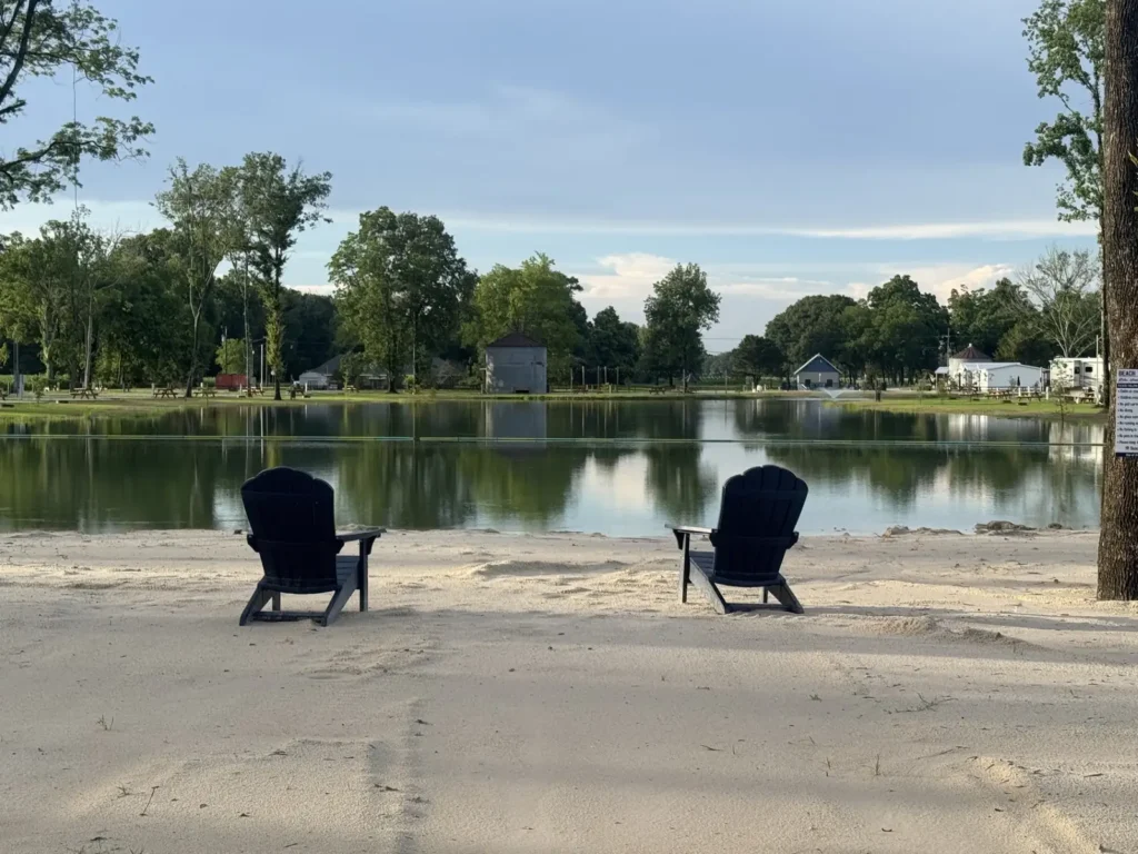 Two black Adirondack chairs sit on a sandy beach facing a calm pond with trees and campground buildings beyond.