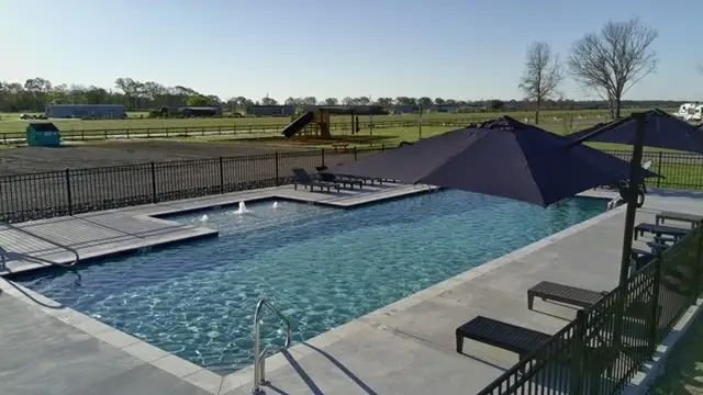 Outdoor swimming pool with tanning ledge fountains, lounge chairs, large blue shade umbrellas, and a playground beyond