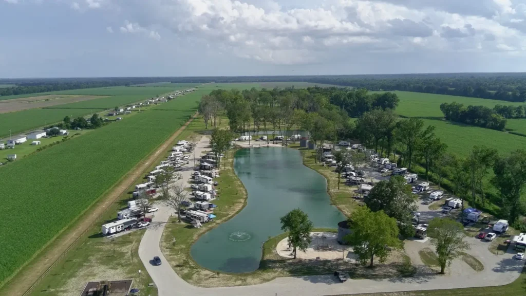 Aerial view of an RV park with dozens of trailers ringing a fountain pond, surrounded by green farm fields.