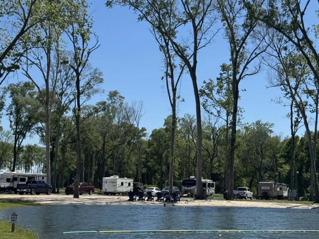 Swim beach on a lake with 'Swim Area Only' sign, Adirondack chairs, parked travel trailers and trucks under tall trees.