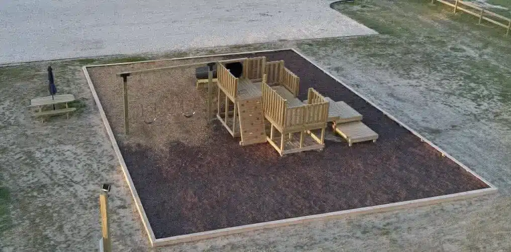 Aerial view of a wooden playground with swings, climbing wall and slide on mulch, plus a picnic table with umbrella.