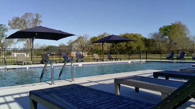 Outdoor swimming pool with handrail steps, navy lounge chairs and shade umbrellas, enclosed by black fencing with trees