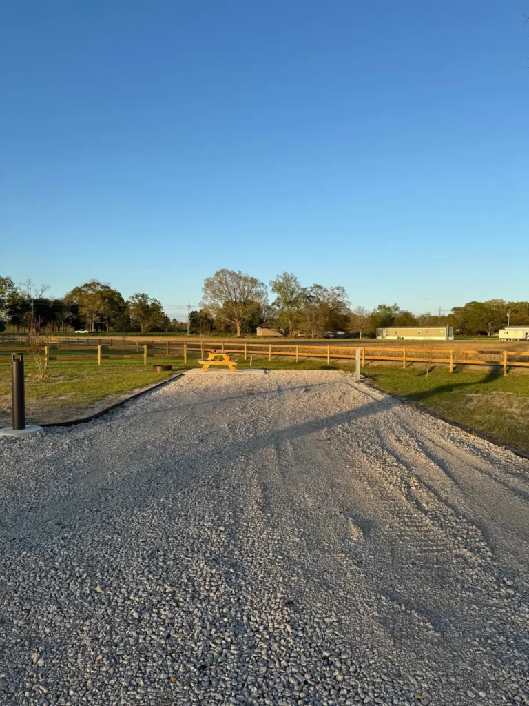 Empty gravel RV site with a black utility pedestal, wooden picnic table, metal fire ring, and wooden fence beyond.