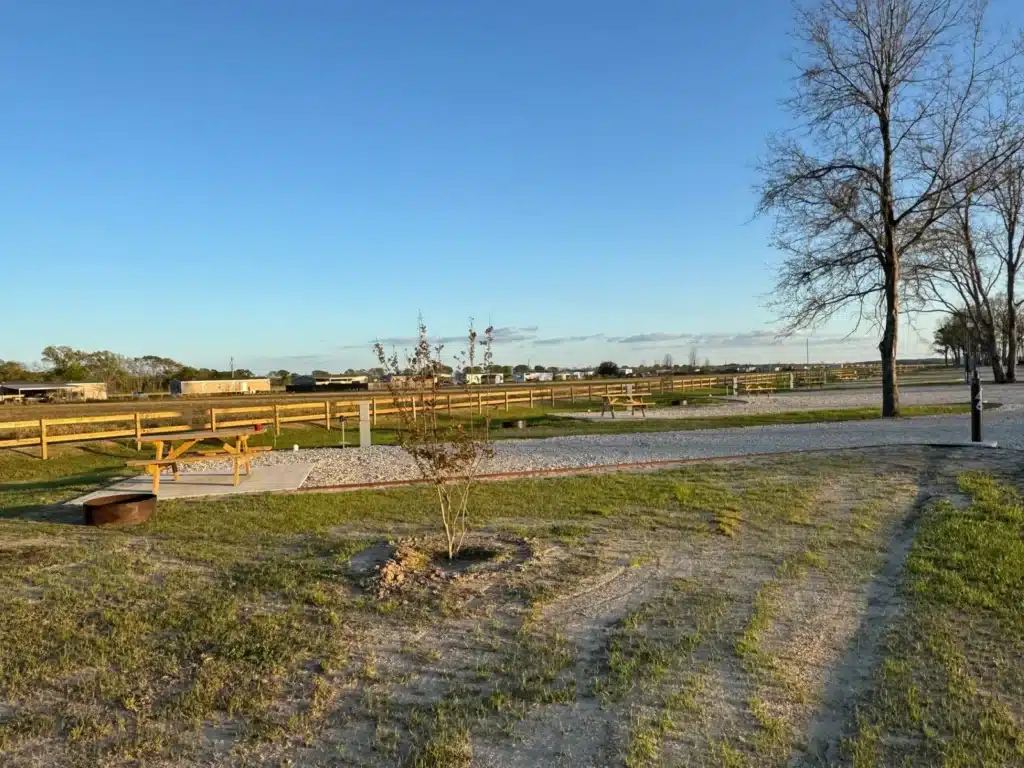 Grassy RV park site with concrete pad, wooden picnic table, metal fire ring, gravel road, split-rail fence and young