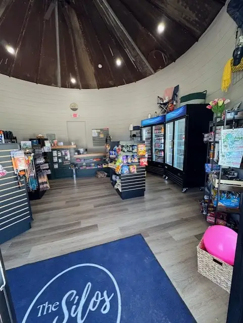 Interior of a round camp store with glass-door drink coolers, snack shelves, merchandise racks, and a navy 'The Silos'