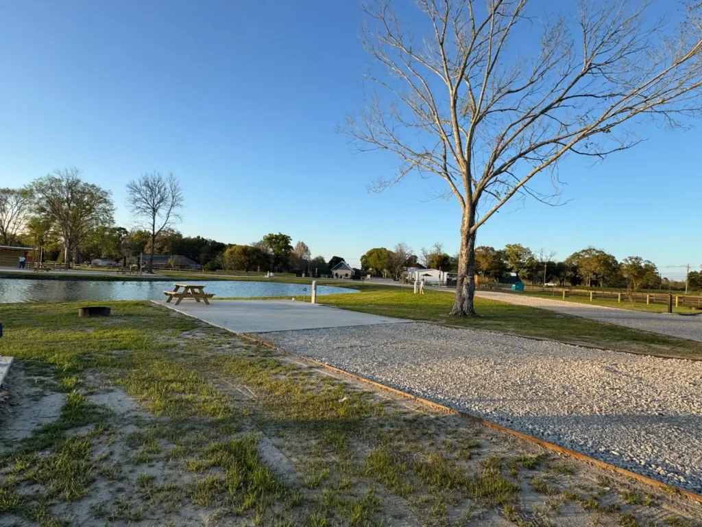 Empty gravel and concrete RV site with wooden picnic table and fire ring beside a pond and bare tree.