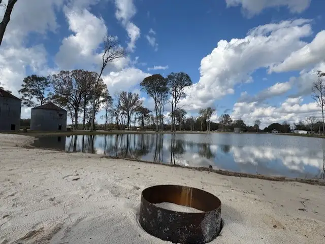 Rusty metal ring fire pit on white sandy beach beside a calm pond with bare trees and grain silos beyond.