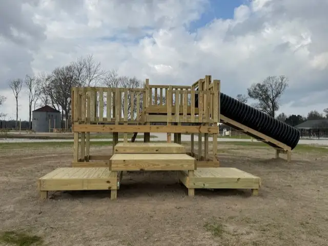 New wooden playground structure with railings, steps, decks and a black corrugated tube slide on bare dirt ground.